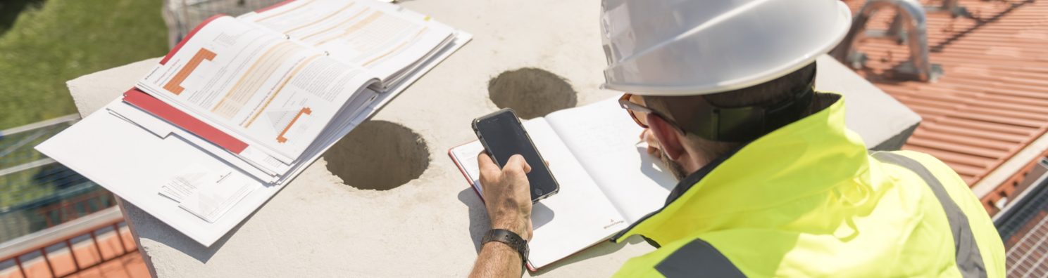 Urban roofer taking notes holding iPhone and writing in an urban location wearing hard hat and safety jacket, Fast Forward Commercial Excellence