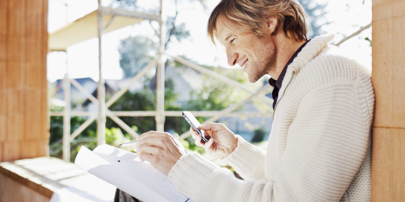 Smiling man with blueprint and mobile phone sitting on window sill at construction site